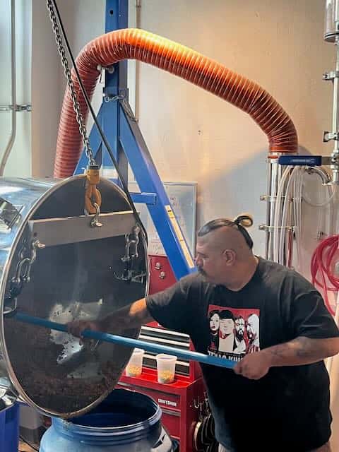 Man stirring brewing ingredients inside a large stainless steel tank at a craft brewery, with ventilation hose above.