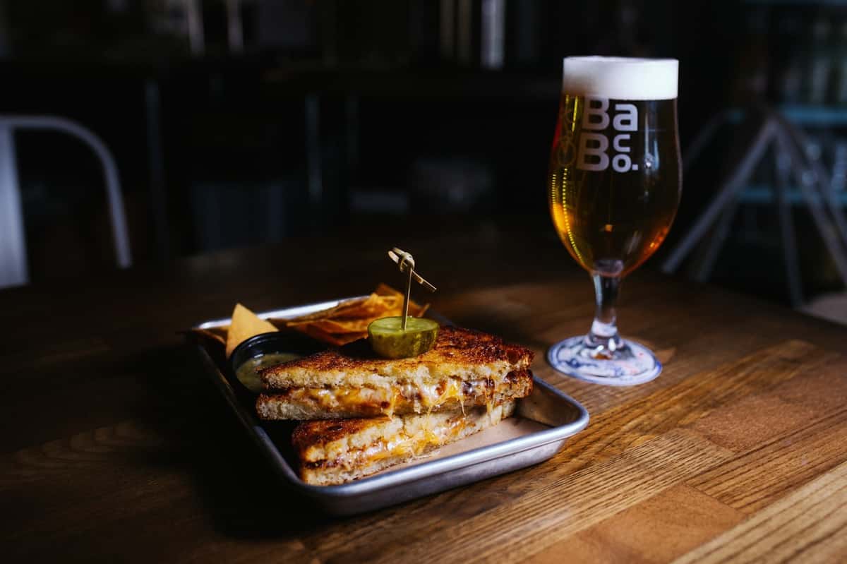 Grilled cheese sandwich with a pickle and chips is served alongside a golden craft beer in a Breakaway Brewing Co. glass.