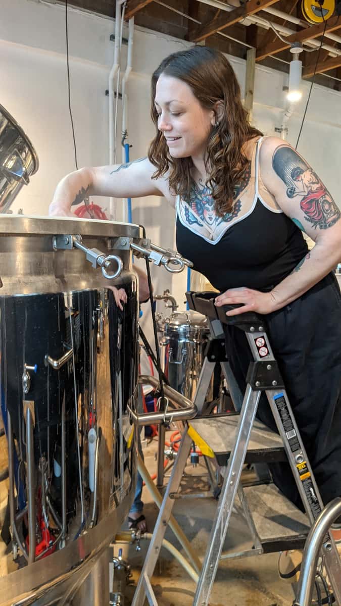 Woman with tattoos standing on a ladder, smiling while working with a large stainless steel brewing tank in a brewery.