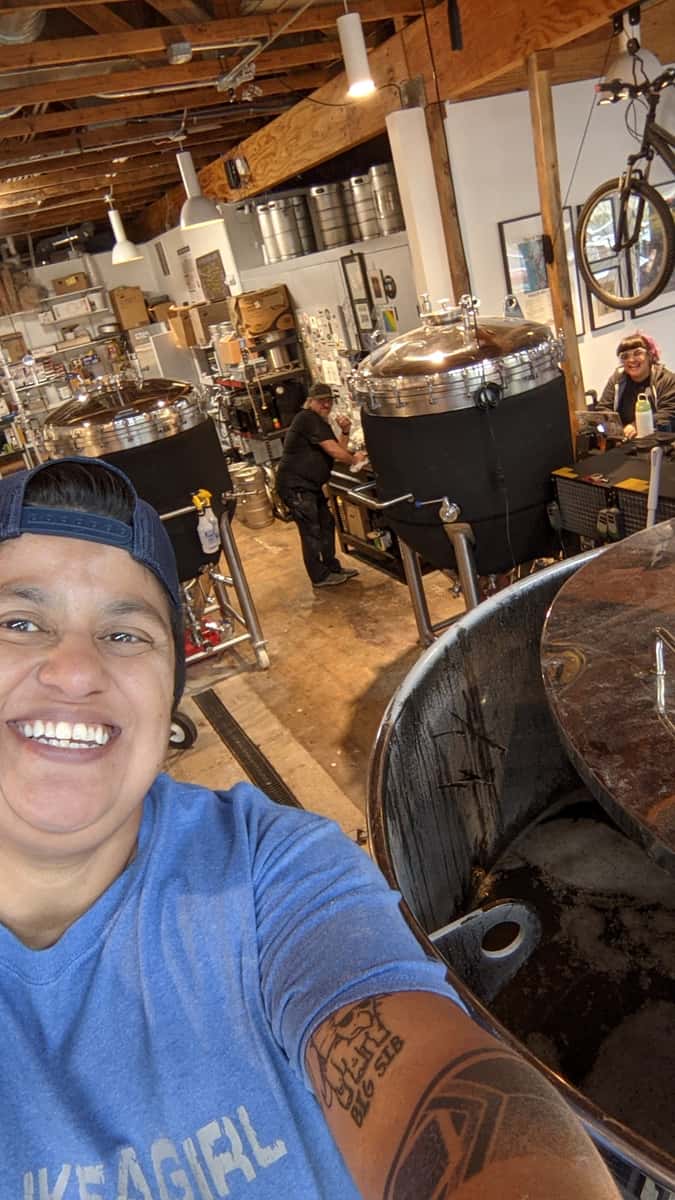 Person taking a selfie in a brewery with large fermentation tanks, smiling workers, and brewing equipment in the background.