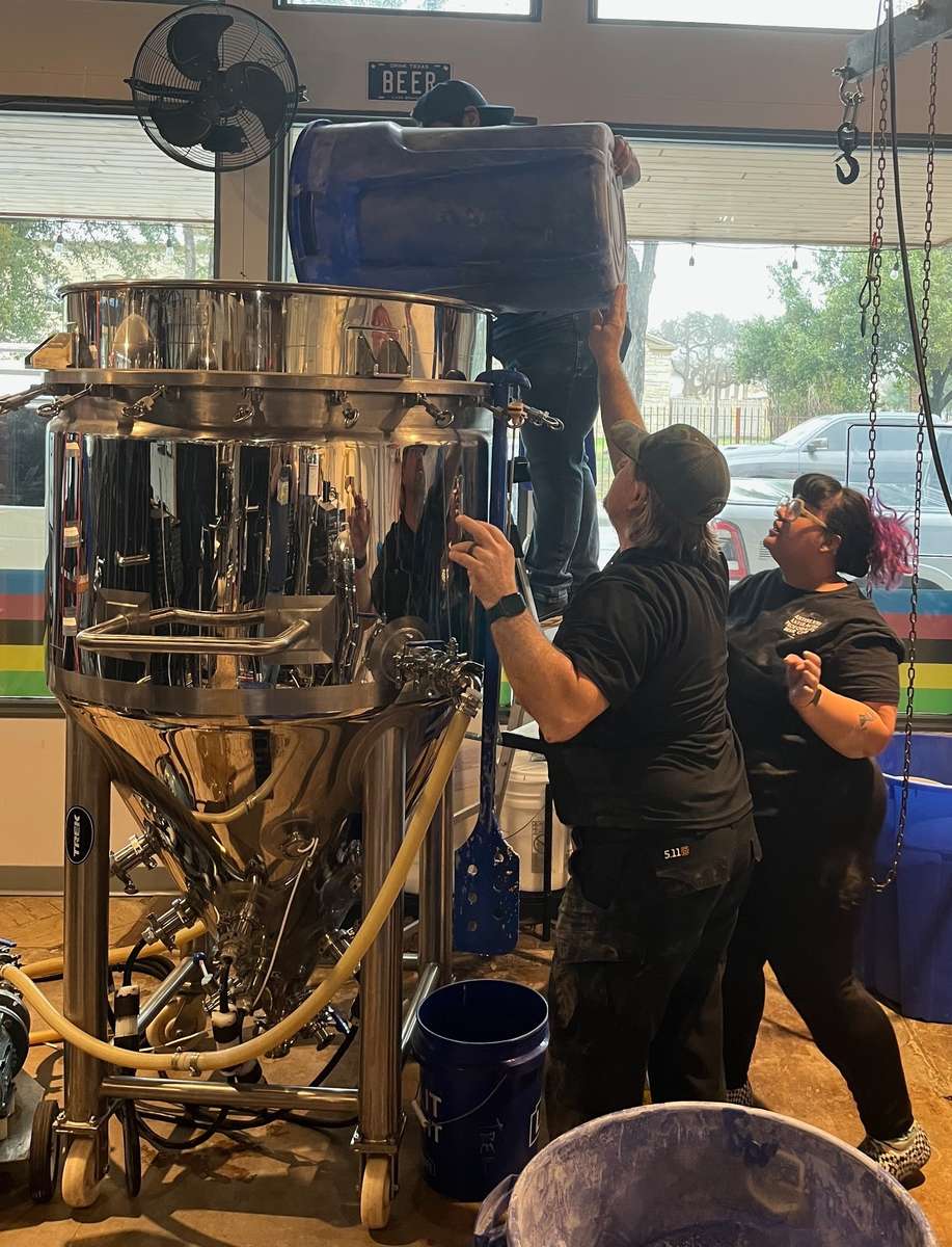 Three brewery workers pour ingredients from a blue container into a large stainless steel fermentation tank near a window.