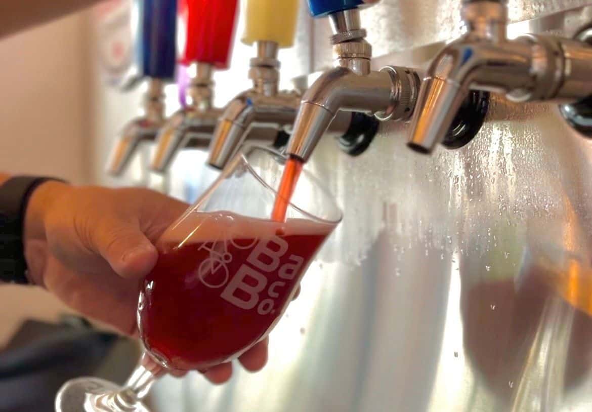 A person fills a branded glass with red craft beer from a row of colorful beer taps mounted on a metal wall.