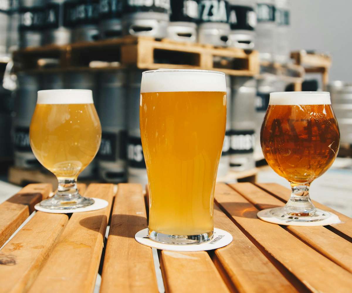 Three craft beers in different shades of amber and gold sit on a wooden table outdoors, with brewery kegs stacked in the background.