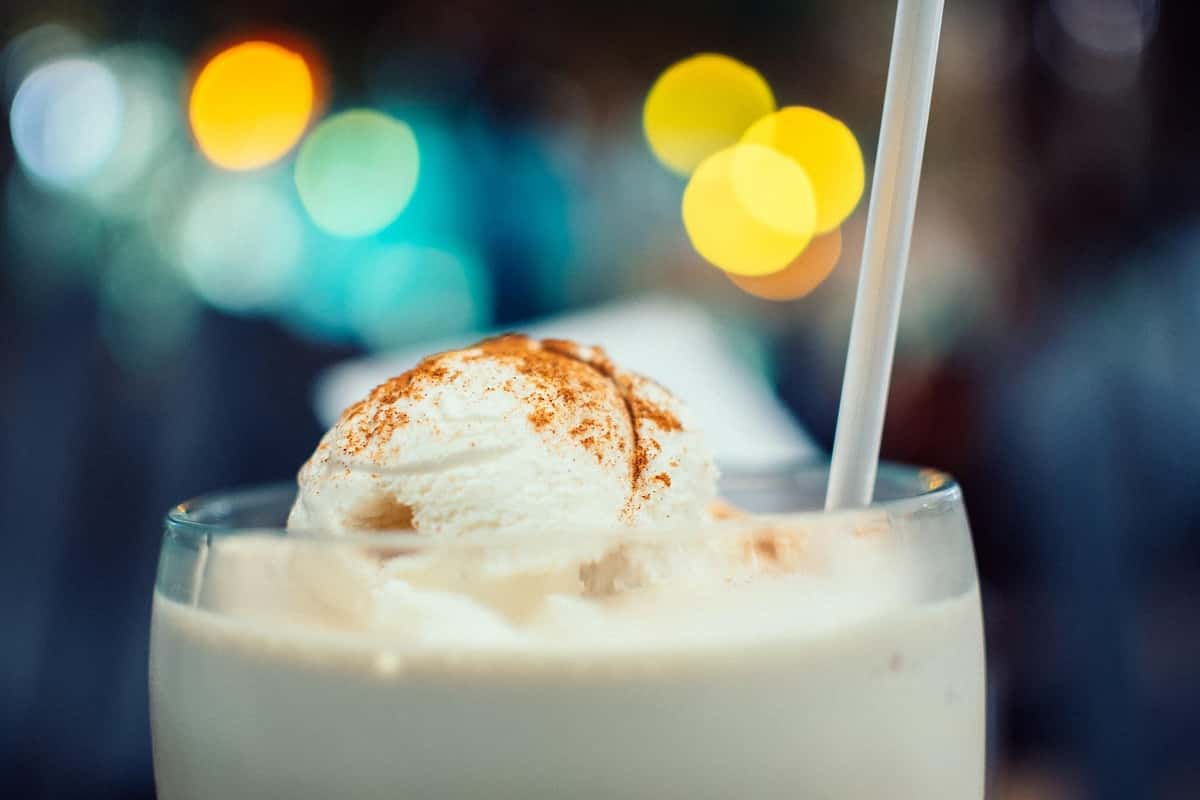 A dessert drink is shown with a scoop of ice cream, a straw, and a dusting of cinnamon. Bokeh lights softly illuminate the background of the image.