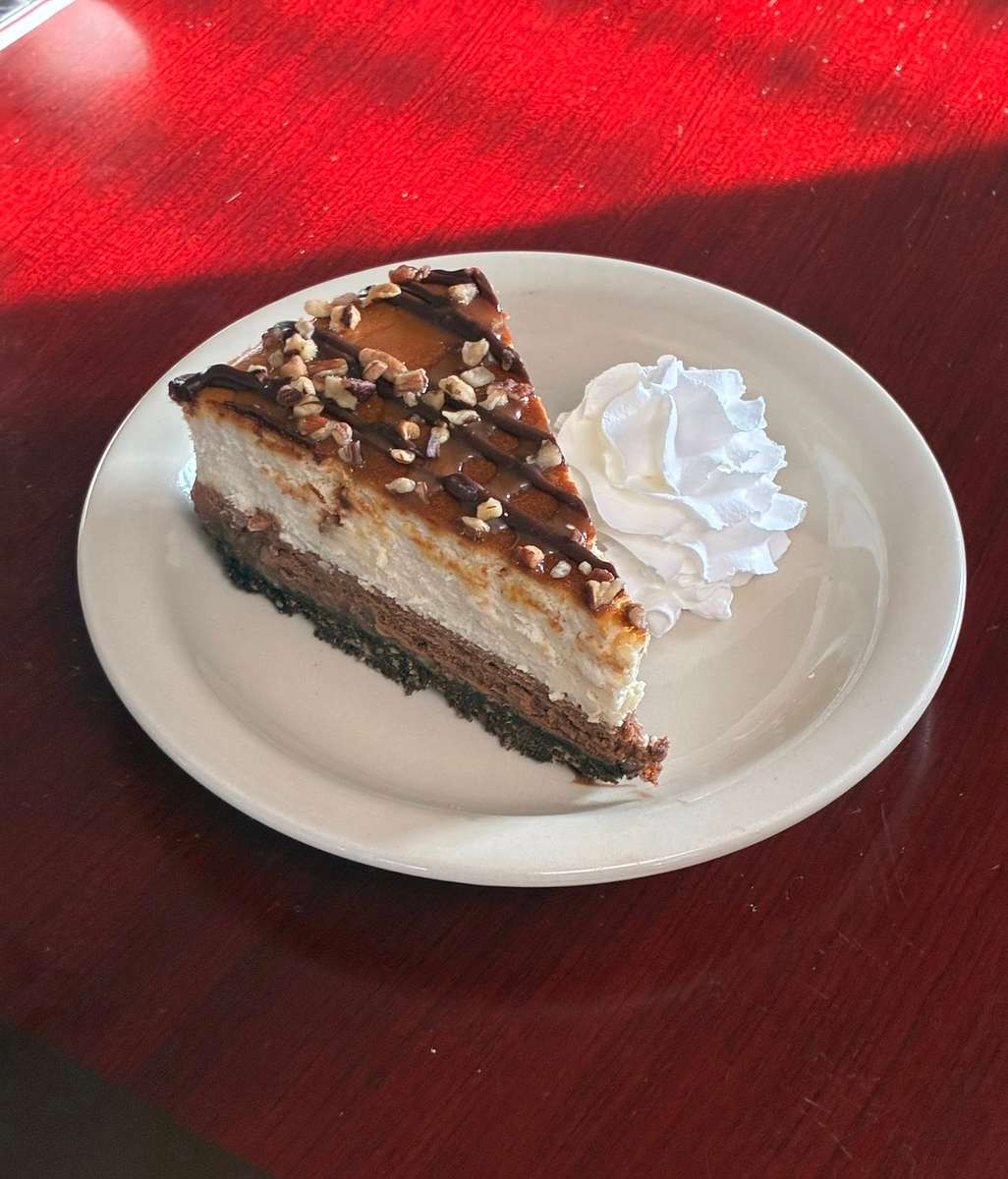 Slice of layered turtle cheesecake topped with chocolate drizzle and chopped pecans, served with a swirl of whipped cream.