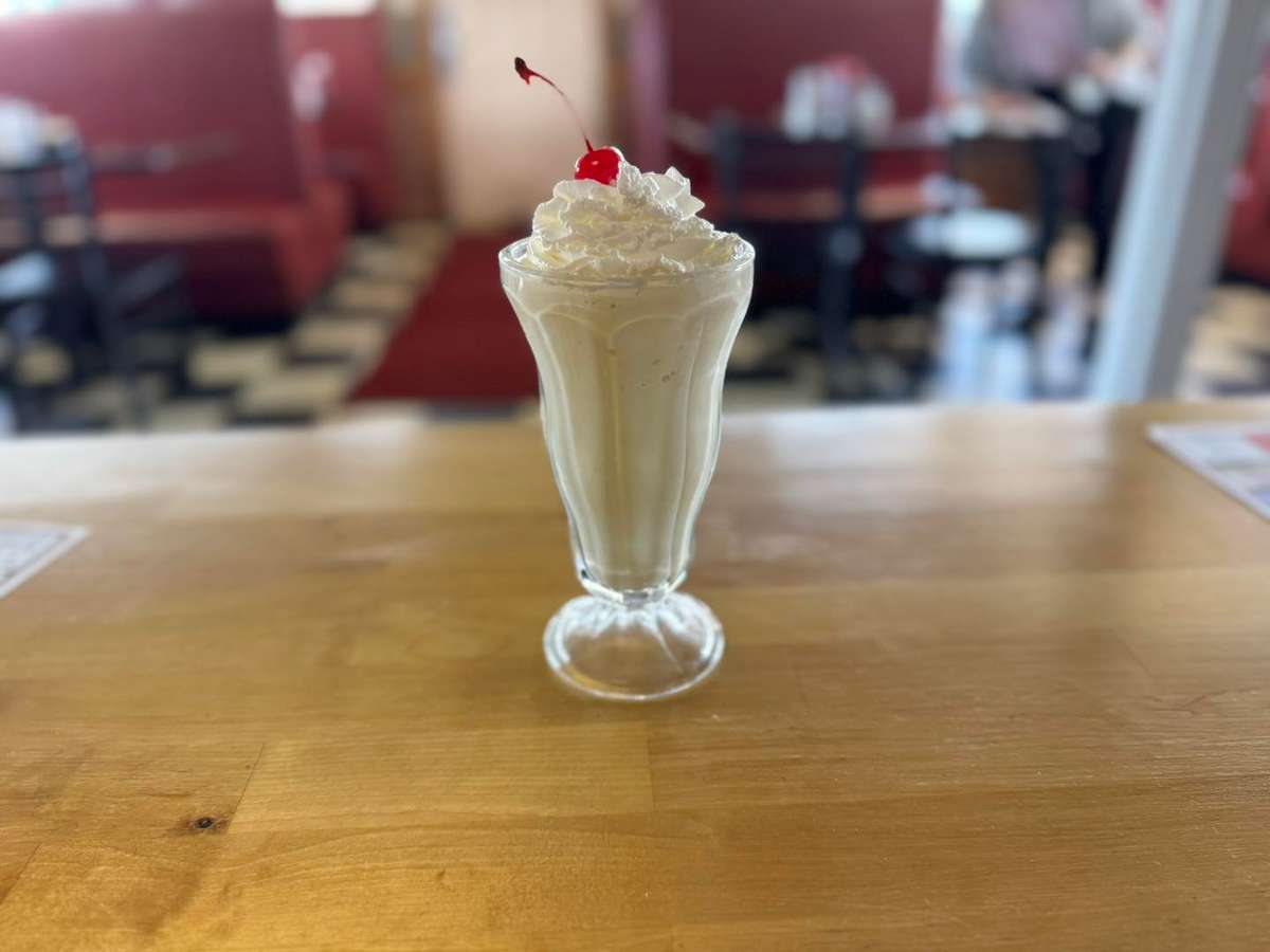 Vanilla milkshake topped with whipped cream and a cherry, served in a tall glass on a wooden counter in a diner setting.
