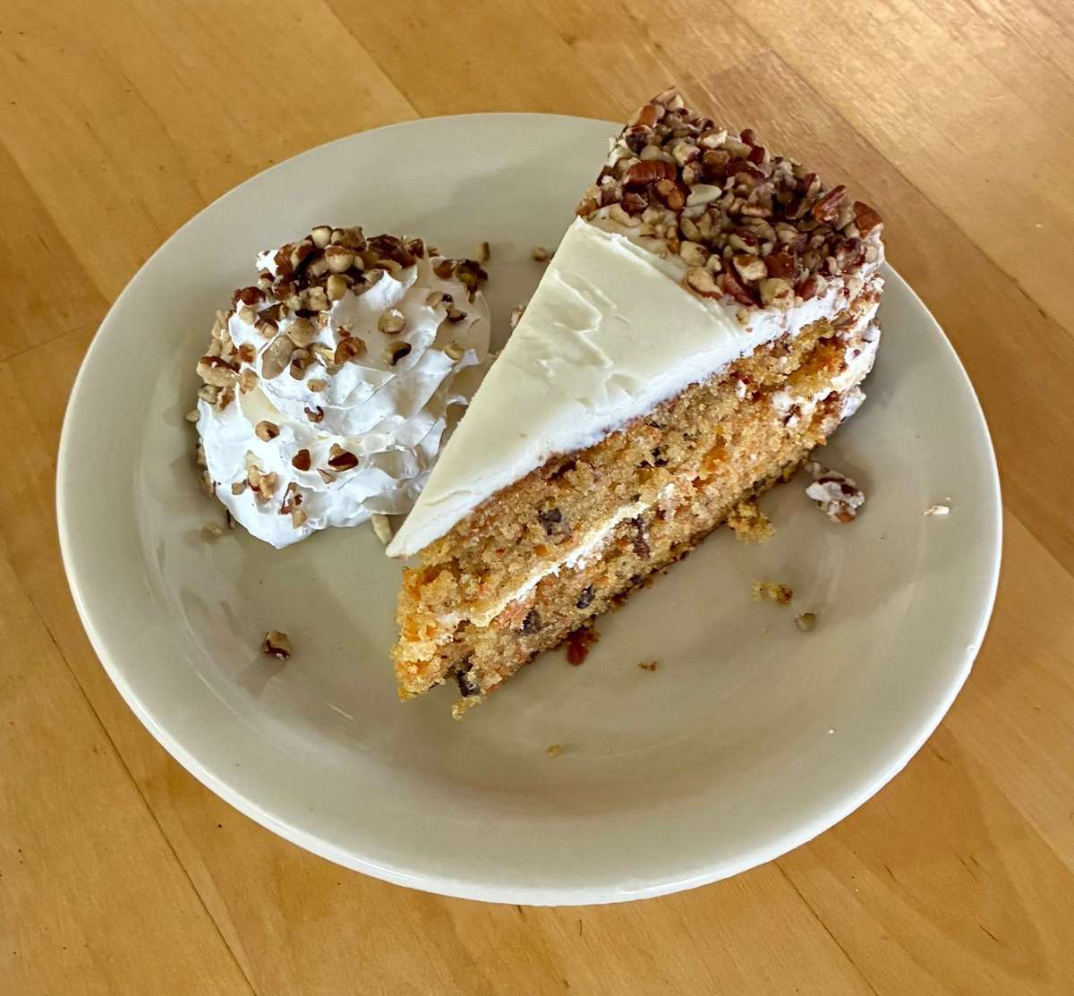 Slice of carrot cake with cream cheese frosting and chopped pecans, served with whipped cream topped with more pecans.