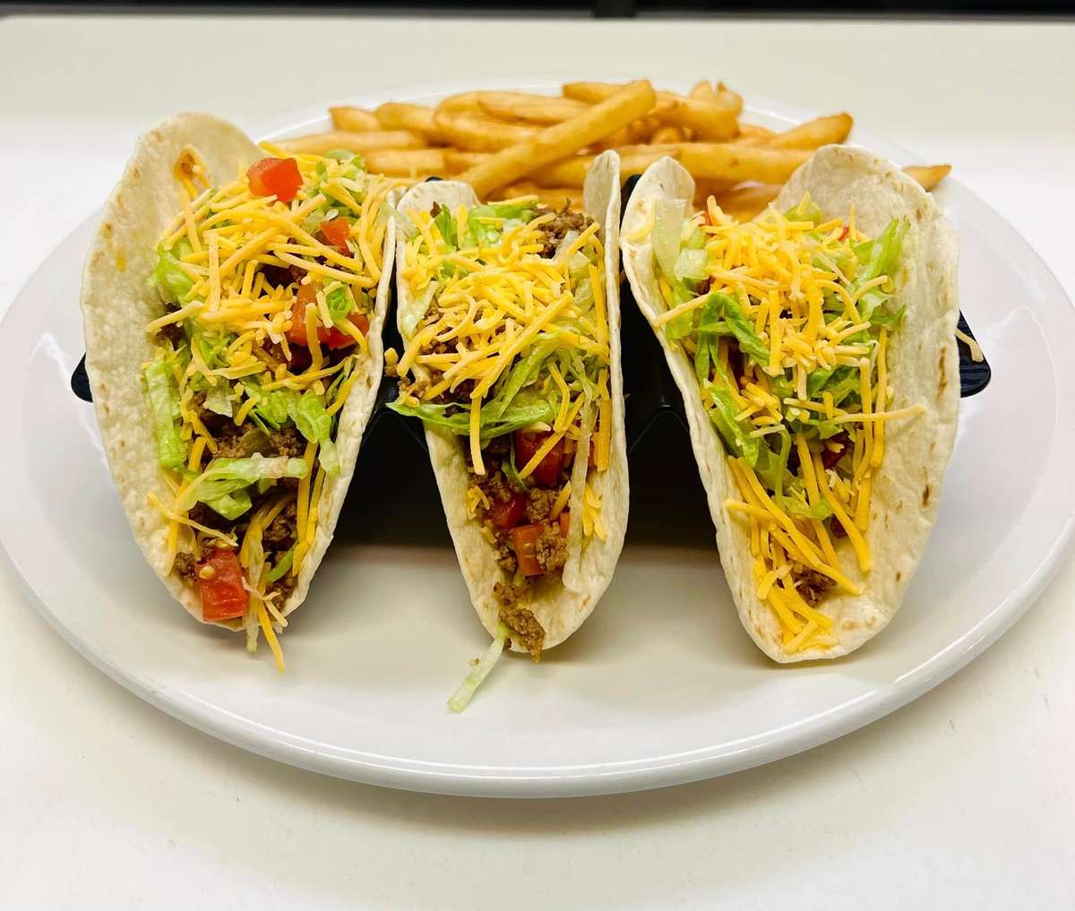 Three soft shell tacos with ground beef, shredded lettuce, diced tomatoes, and cheddar cheese, served with French fries.