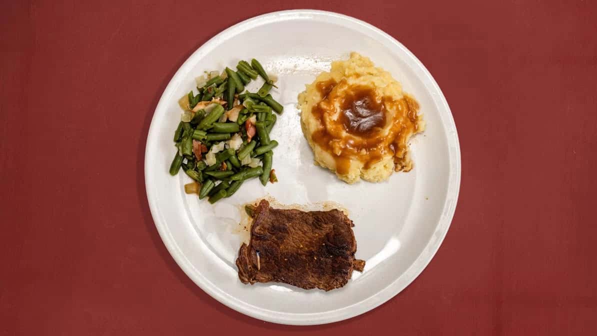 Pan-seared beef steak served with mashed potatoes topped with brown gravy and a side of green beans with onions and bacon.