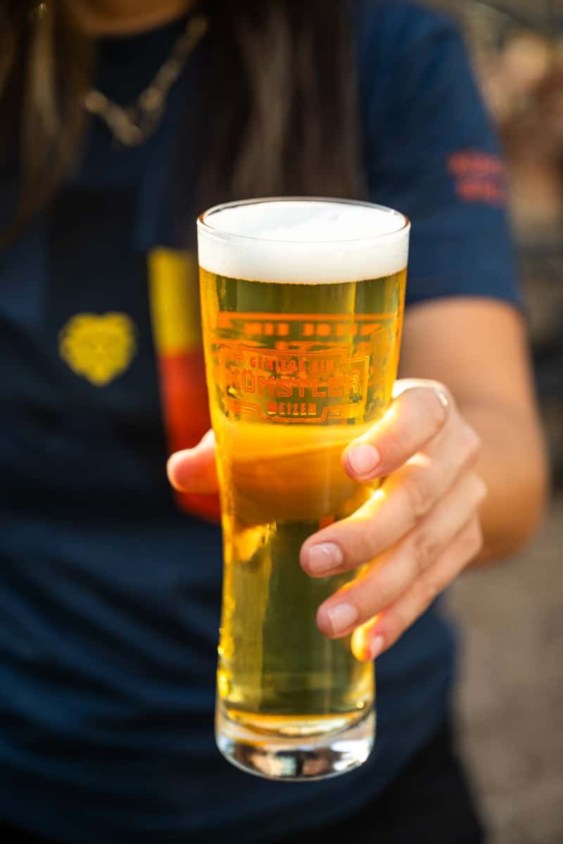 Close-up of a golden Weizen beer in a branded Künstler Brewing glass, freshly poured with a frothy head, held outdoors by a staff member in a brewery t-shirt.