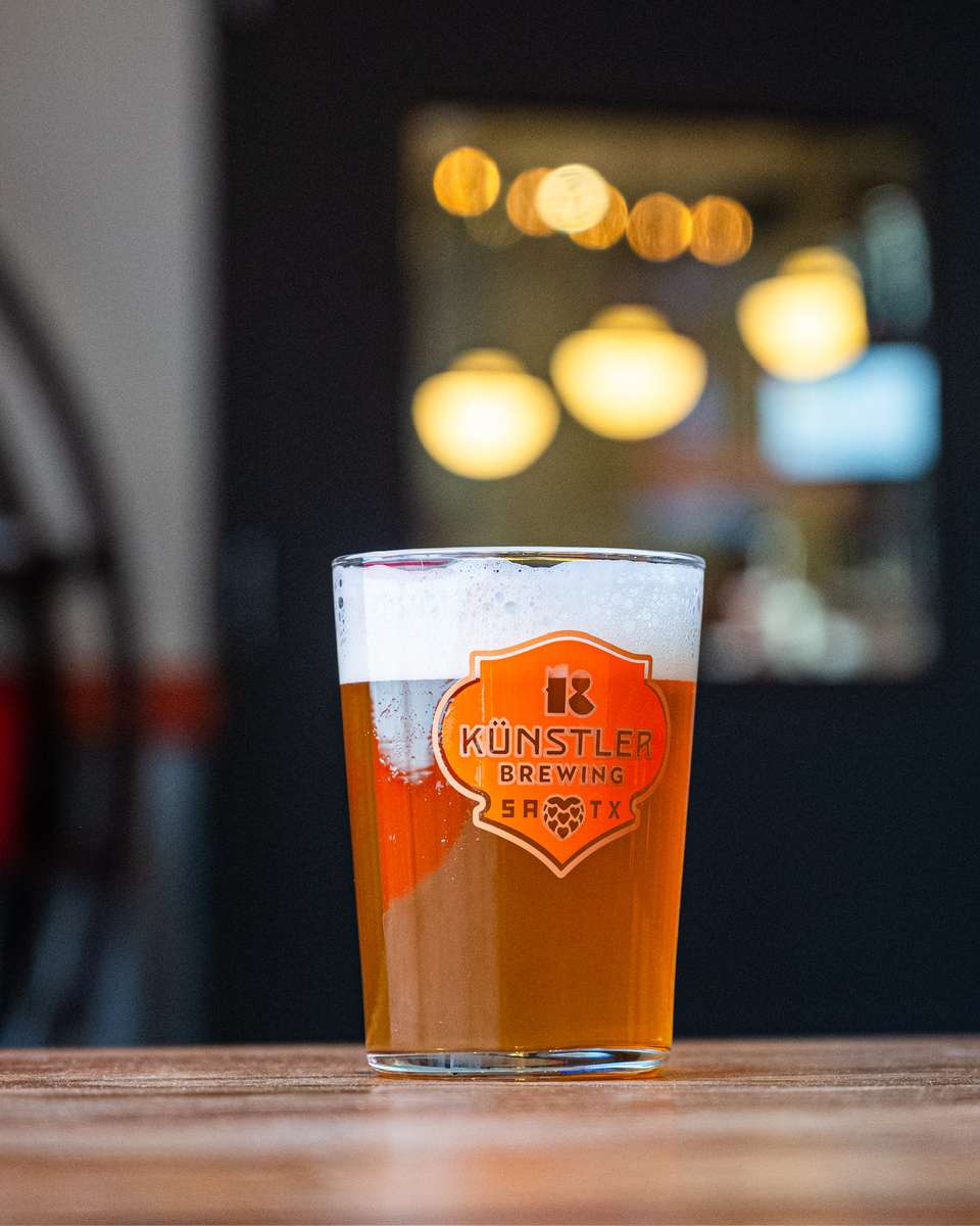 Pint of Künstler Brewing beer on a wooden table, with bokeh lights and a cozy brewery interior softly glowing in the background.