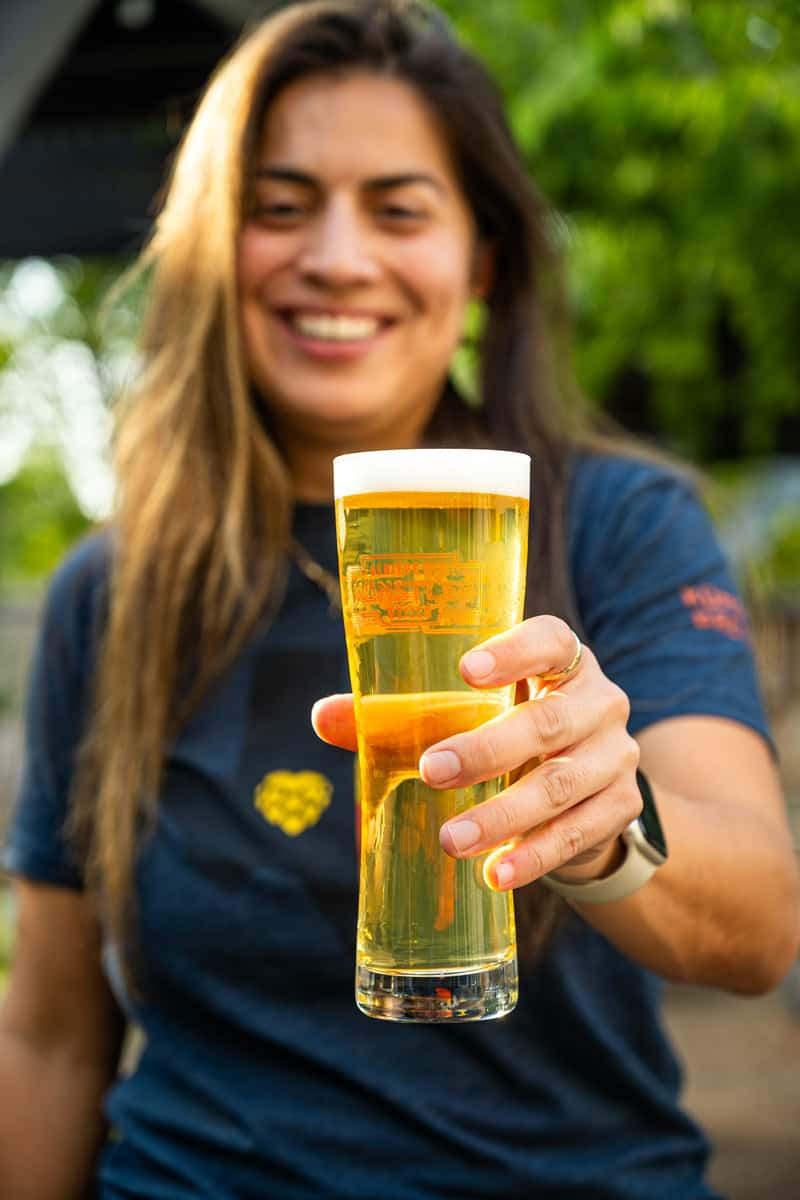 Smiling woman holding a tall glass of golden craft beer with a frothy head, served at Künstler Brewing, in a vibrant outdoor setting.
