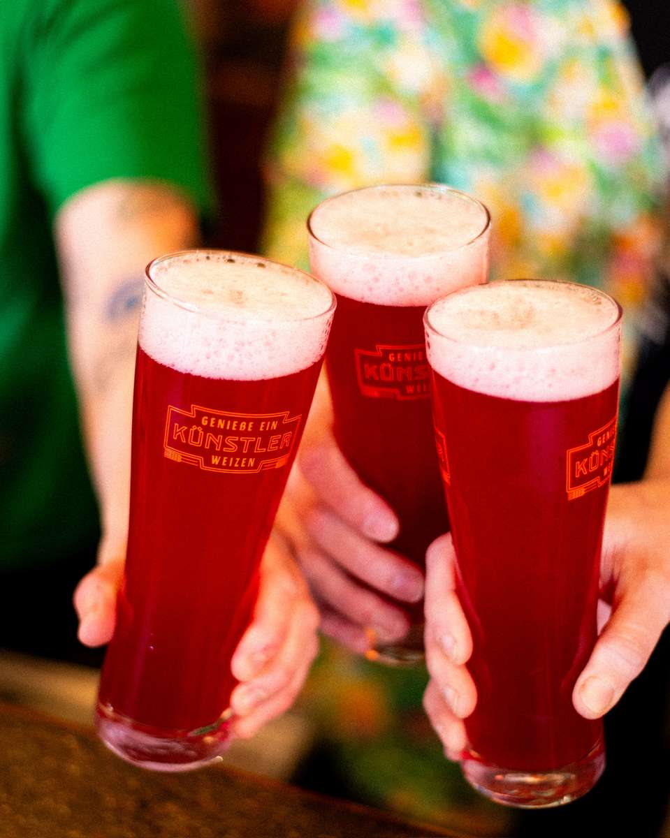 Close-up of three tall weizen glasses filled with ruby-hued fruit beer from Künstler Brewing, topped with a frothy head.