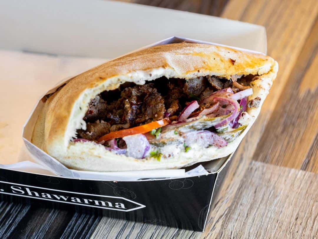 Beef shawarma stuffed in pita bread with onions, tomatoes, pickles, and garlic sauce, served in branded packaging.