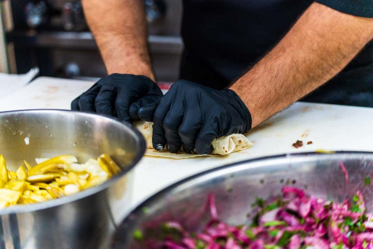 This image shows a person wearing black gloves carefully rolling a shawarma wrap on a white preparation surface. In the foreground, there are stainless steel mixing bowls — one containing pickled vegetables (likely pickles or pepperoncini) and the other filled with a chopped salad of red onions and herbs, likely parsley. The person appears to be in the process of assembling and wrapping the shawarma for serving.