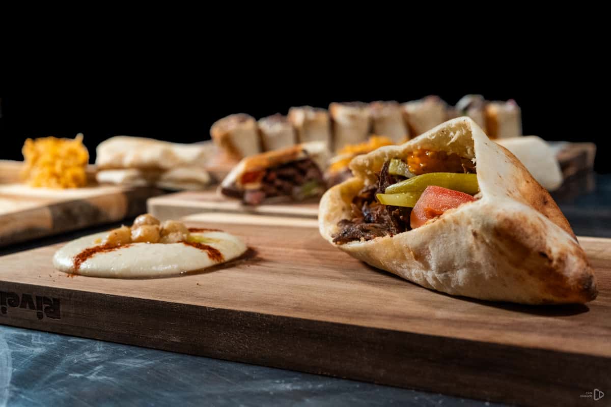 Stuffed pita with beef shawarma, tomatoes, and pickles served on a wooden board with hummus and assorted wraps in the background.