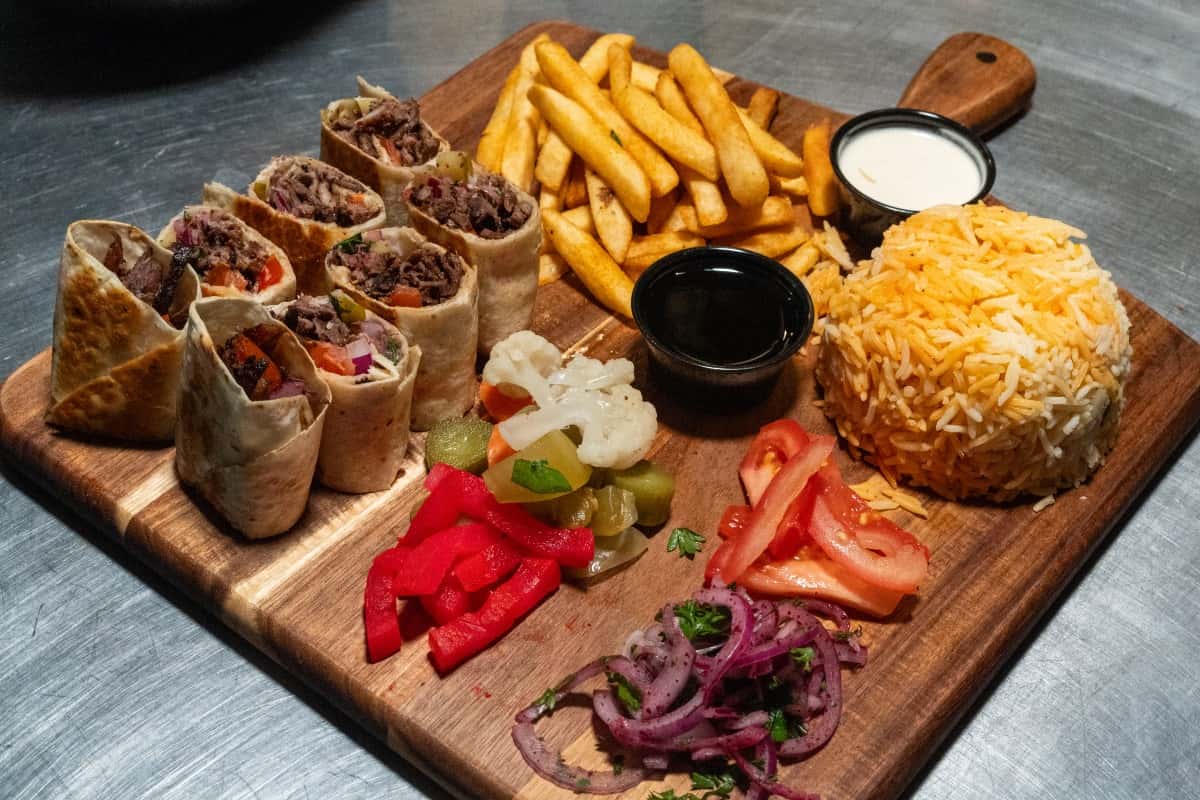 Beef shawarma wrap slices served with fries, rice, pickled vegetables, tomatoes, onions, and dipping sauces on a wooden board.