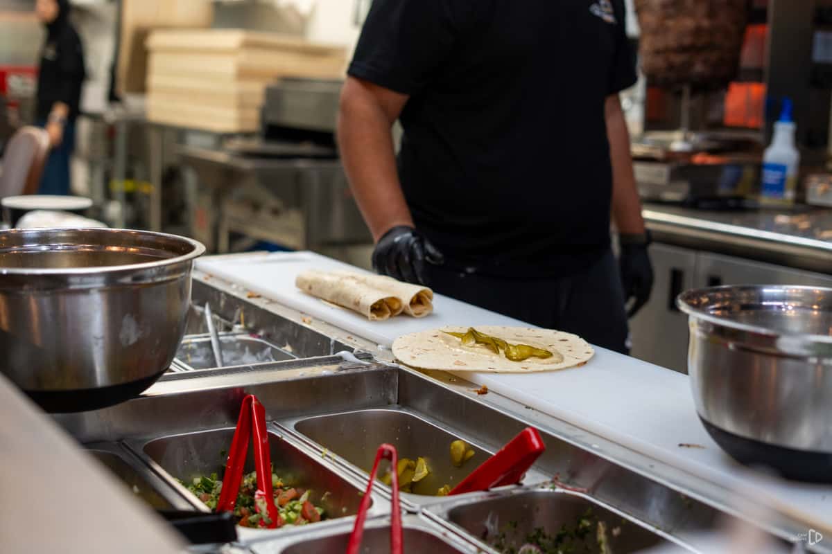 Shawarma prep station with tortillas on a cutting board, pickles added, and staff working among stainless steel ingredient bins.