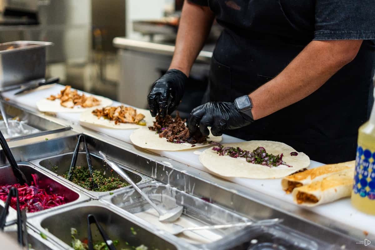 Worker assembling shawarma wraps on a prep counter with toppings like meat, parsley, and pickled vegetables from a salad bar.
