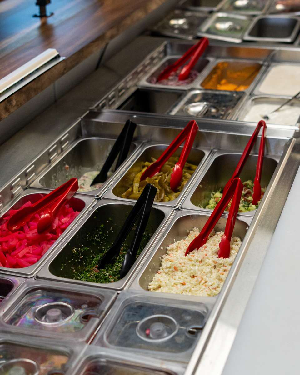 Salad bar with various Middle Eastern toppings like pickled turnips, tabbouleh, jalapeños, coleslaw, and sauces with tongs.