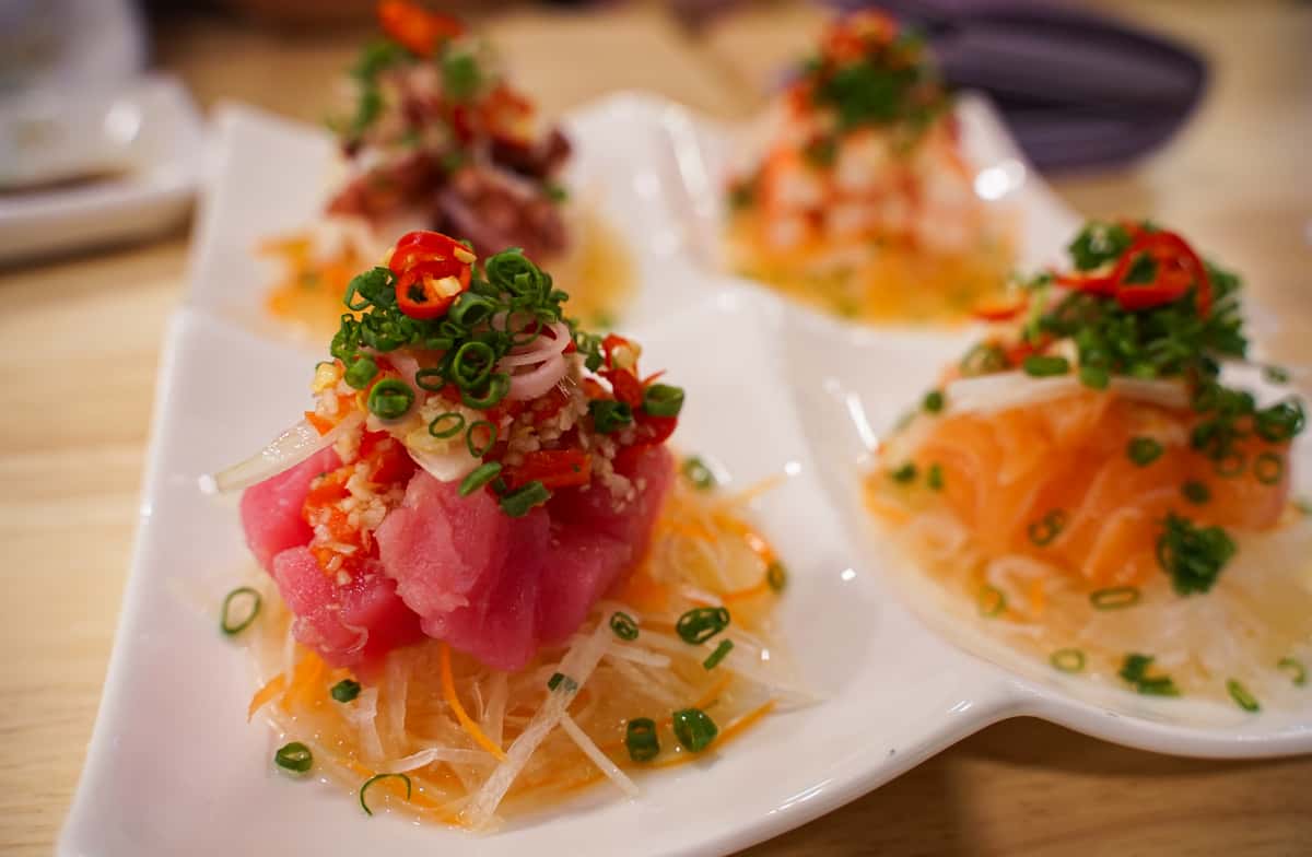 A close-up of three small, elegant appetizers served on a white wavy plate. Each portion consists of finely diced tuna or salmon tartare topped with fresh microgreens.