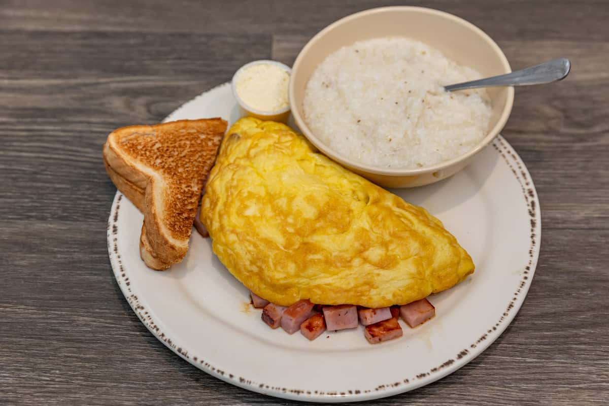Hearty Southern-style breakfast plate featuring a fluffy ham-stuffed omelet, creamy white grits with a spoon, buttered toast, and a side of whipped butter — a classic comfort food combo ideal for American diner menus.