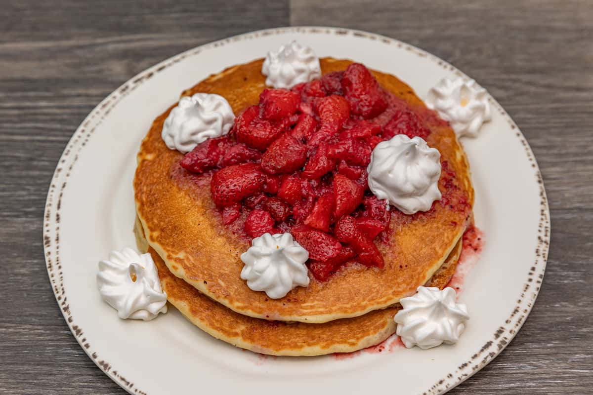 Stack of fluffy golden pancakes topped with a generous serving of syrupy strawberries and surrounded by dollops of whipped cream, served on a rustic ceramic plate — a classic American breakfast dish perfect for brunch menus.