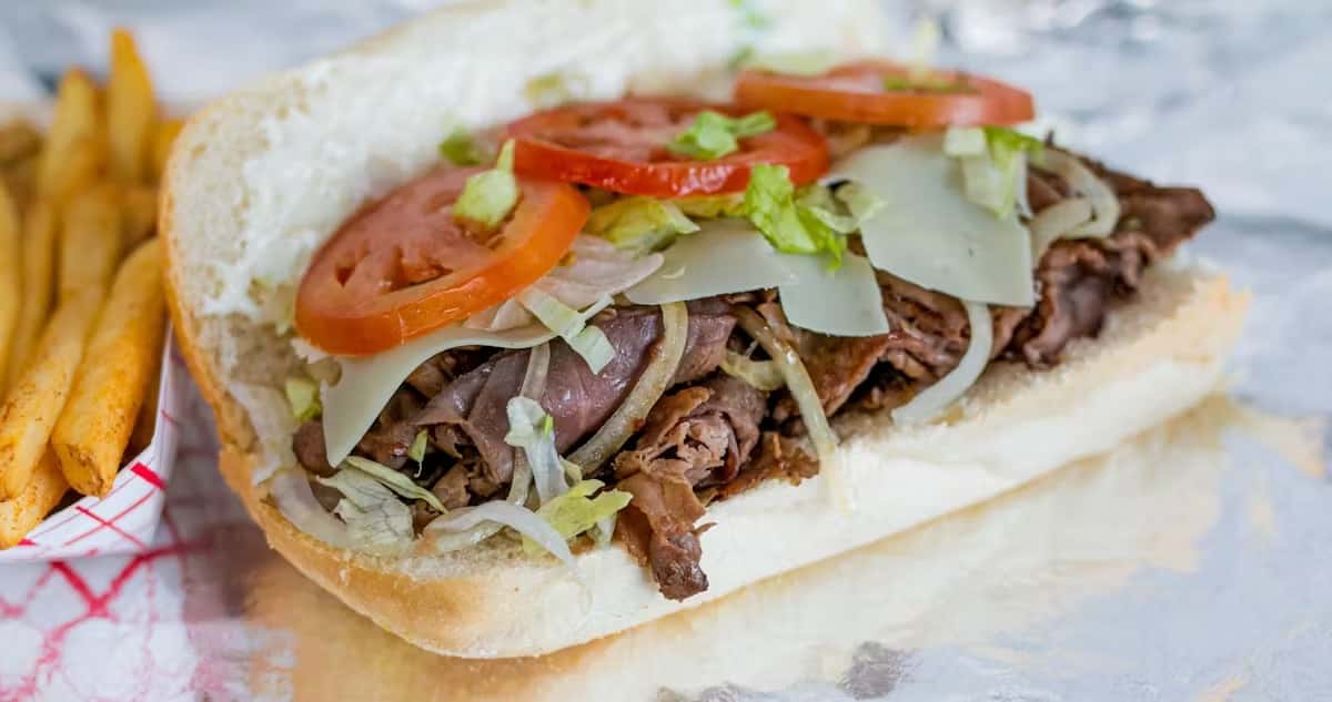 A steak and cheese sub with tomatoes, lettuce, and onions, alongside a side of fries, a featured item at Steak N' Cheese Please.