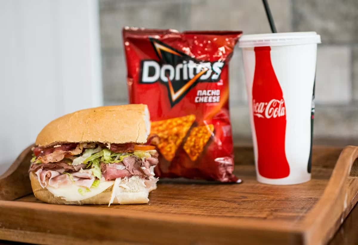 A sandwich, Doritos, and Coca-Cola drink sit on a wooden tray, presenting a meal option from Steak N' Cheese Please.