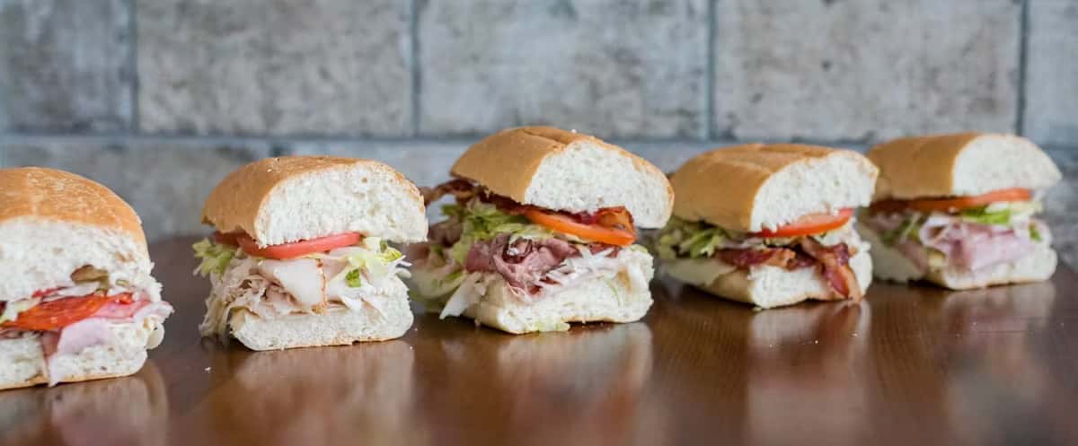 Five halved sub sandwiches with various fillings are lined up on a reflective surface at Steak N' Cheese Please.