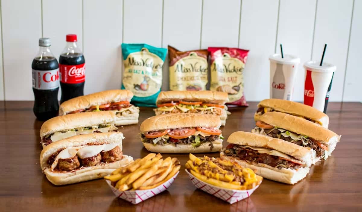 Variety of sandwiches, fries, chips, and drinks arranged on a table at Steak N' Cheese Please.