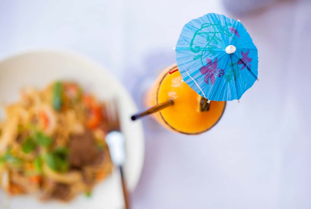 Overhead shot of an orange drink with a blue paper umbrella, next to a blurred plate of food.