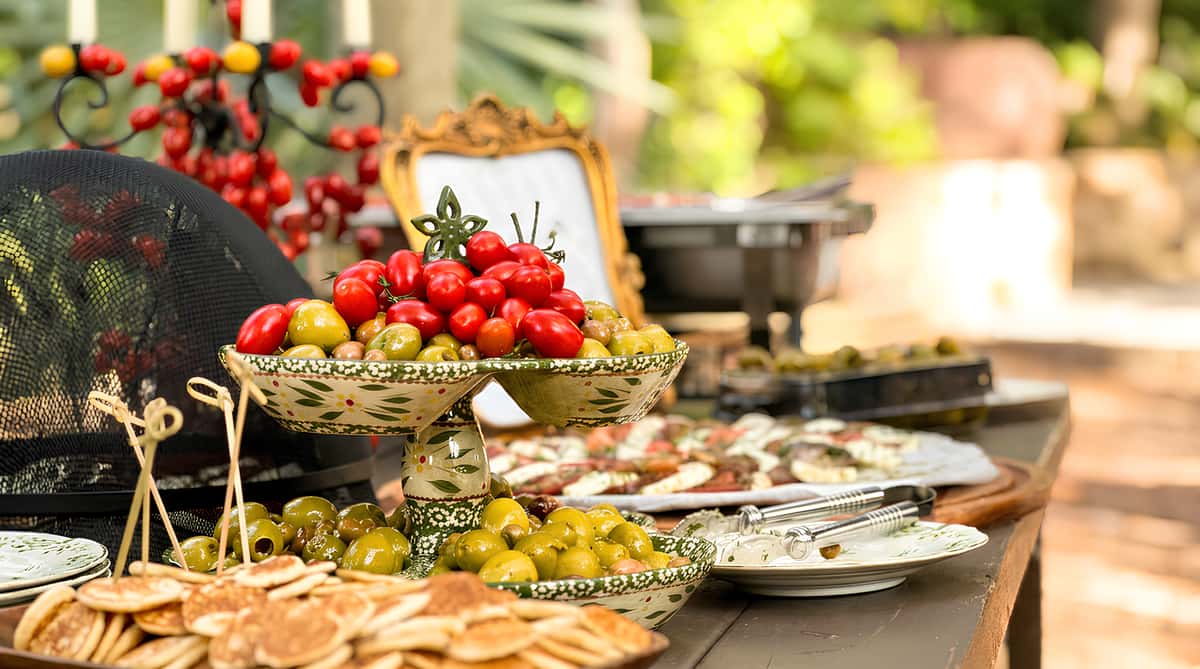Buffet table with a tiered bowl of cherry tomatoes and olives, a dish of small pancakes, and a person's hand reaching for food.