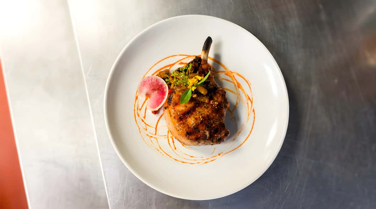 Grilled pork chop with caramelized onions, fresh herbs, and a radish slice on a white plate with red sauce drizzles.
