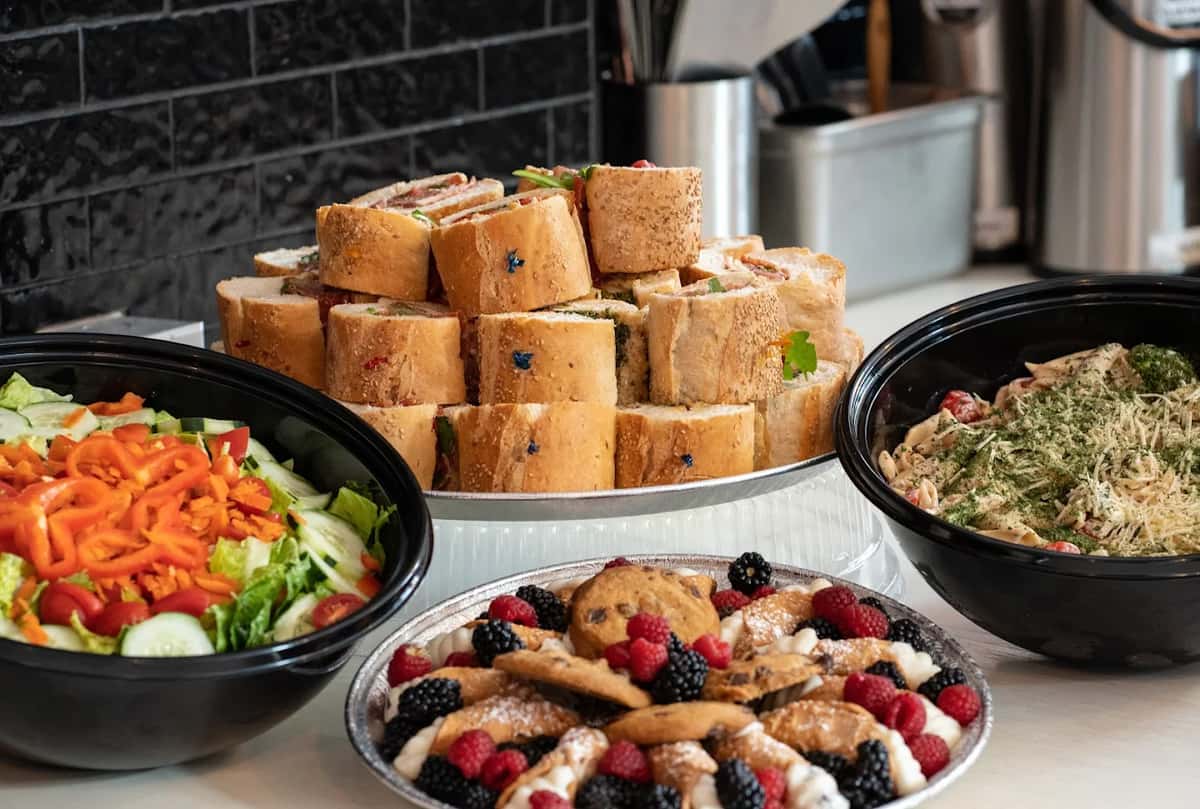 Italian catering platter featuring mini hoagie sandwiches, fresh garden salad, creamy pesto pasta, and assorted Italian desserts with berries and cookies.