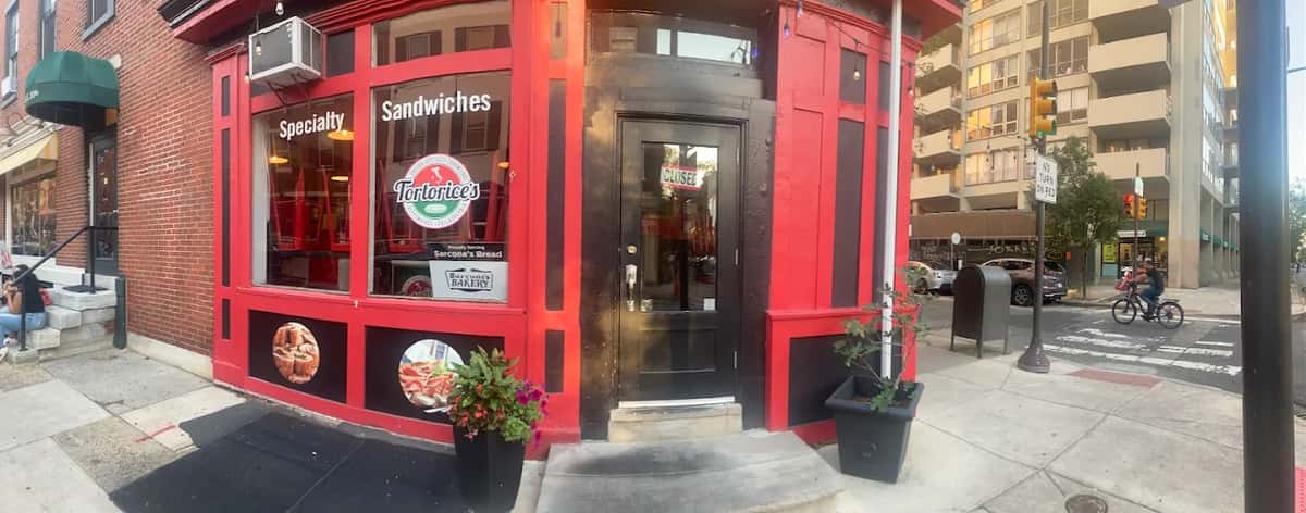 Street view of Tortorice's Italian Specialty Sandwiches in Philadelphia, showcasing its bold red and black exterior, Sarcone's Bakery partnership signage, and large windows promoting specialty sandwiches near a bustling city intersection.