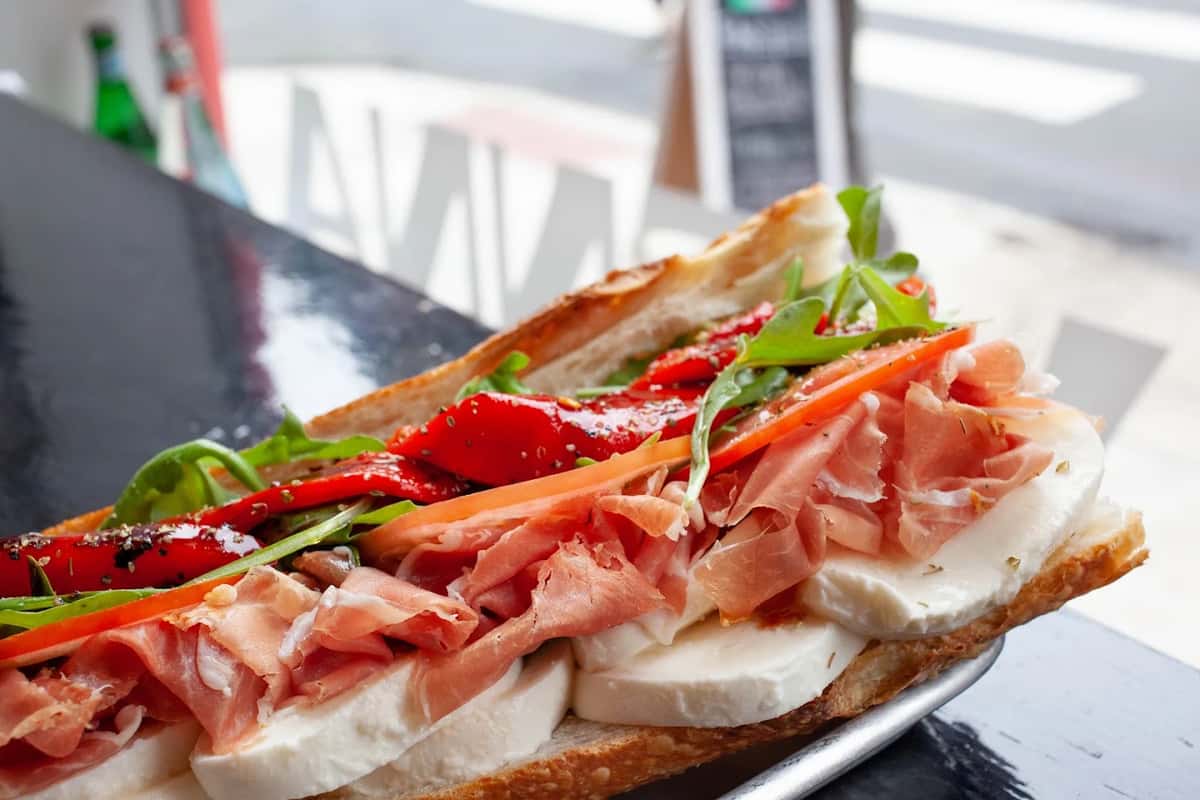 Classic Italian sandwich with layers of fresh mozzarella, prosciutto, arugula, and roasted red peppers on crispy Italian bread, displayed near a deli window.