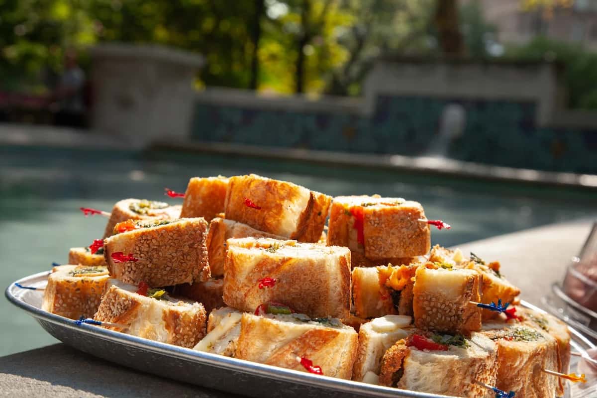 Outdoor catering tray of mini Italian-style sandwiches with assorted fillings, served near a decorative fountain for a picnic or party vibe.