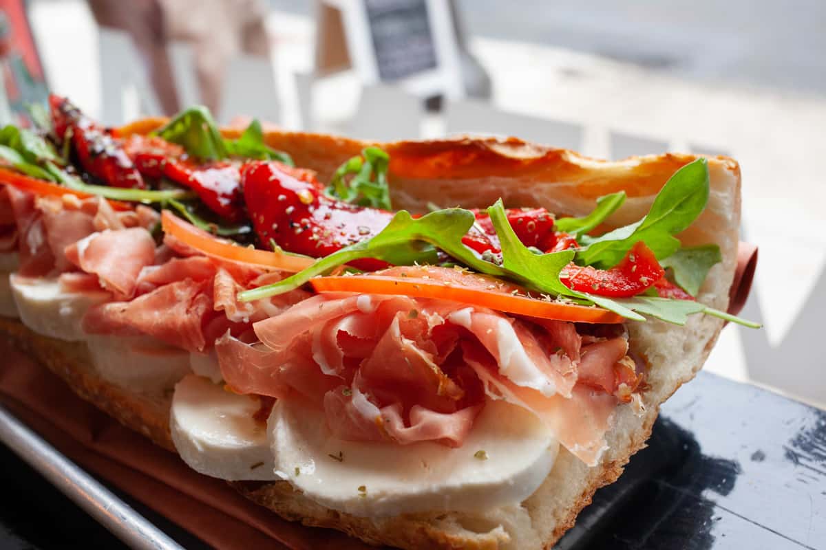 Italian sandwich packed with creamy mozzarella, thinly sliced prosciutto, roasted red peppers, arugula, and tomato on fresh Italian bread, served in a sunlit deli setting.