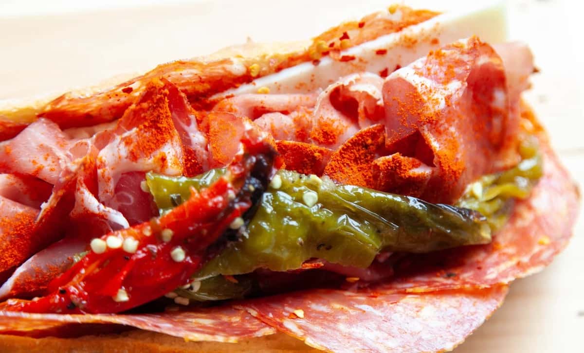 Bold close-up of a spicy Italian sandwich loaded with capicola, salami, roasted red and green peppers, and sprinkled with red chili flakes.