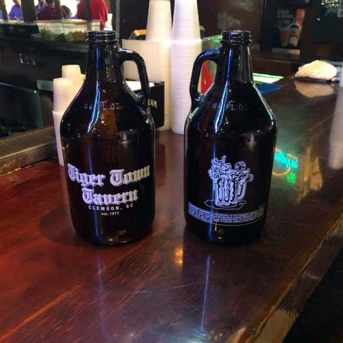 Two dark glass growlers filled with craft beer sitting on a polished wooden bar top. Each growler features a brewery-style logo, while the warm lighting and blurred background create a cozy, laid-back taproom atmosphere perfect for enjoying locally brewed drinks.