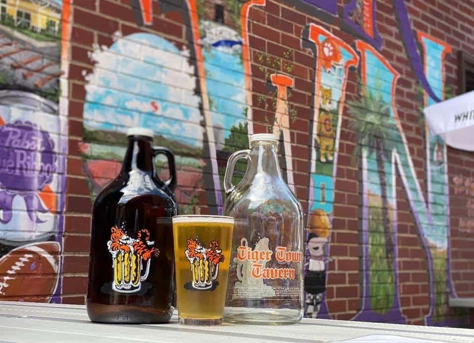 A rustic brewery-style setup featuring craft beer bottles, a filled pint glass, and a clear growler displayed in front of a colorful graffiti wall. The urban backdrop and warm tones create a trendy, laid-back atmosphere that highlights local beer culture and casual social vibes.