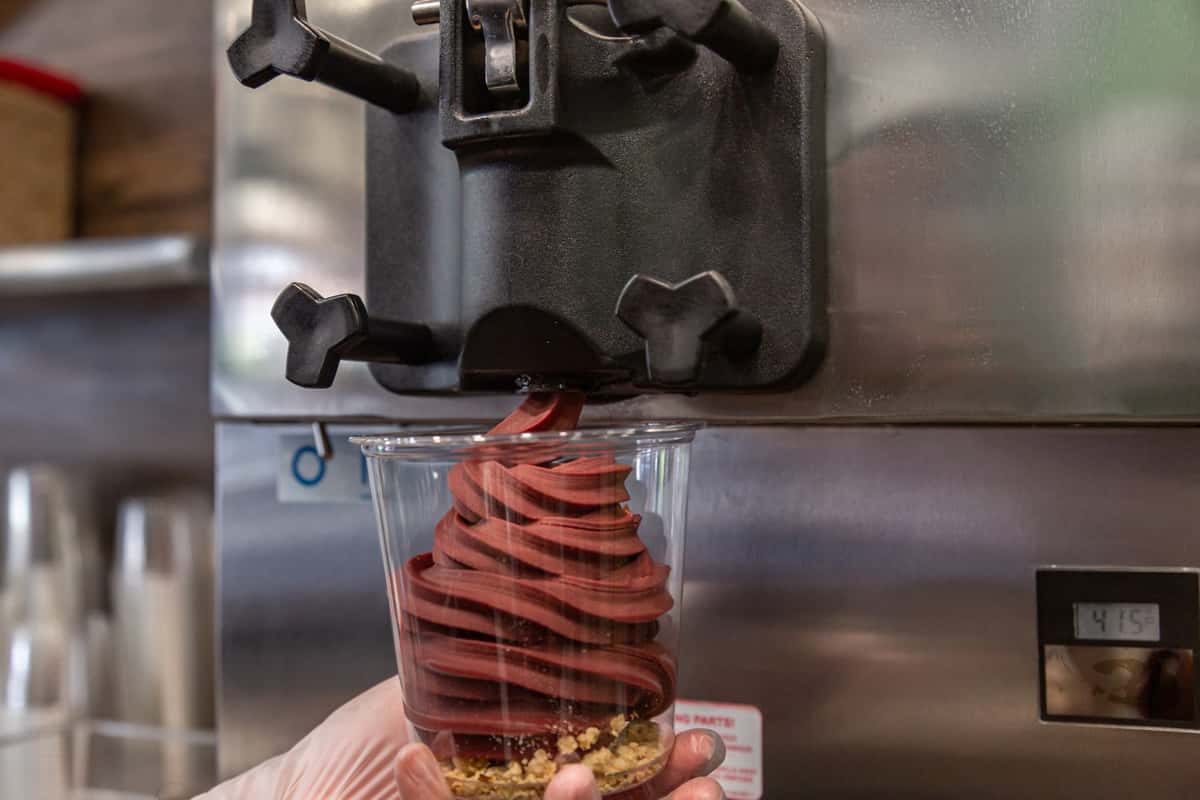 Close-up of chocolate soft serve frozen yogurt being dispensed into a clear plastic cup over a crunchy granola base, served fresh from a commercial frozen yogurt machine.