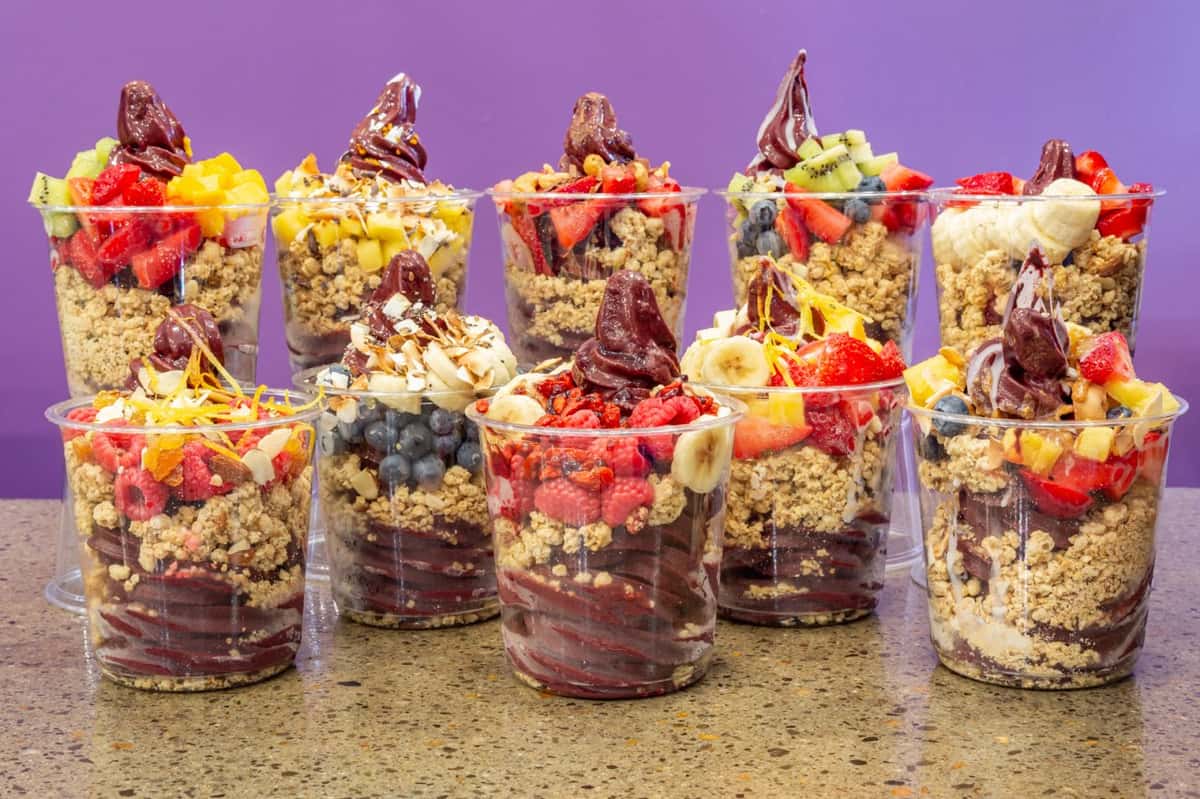 Assorted acai bowls in clear cups lined up on a countertop, each layered with creamy acai blend, crunchy granola, and vibrant fresh fruit toppings like banana, kiwi, strawberries, blueberries, mango, and shredded coconut, against a bright purple background.