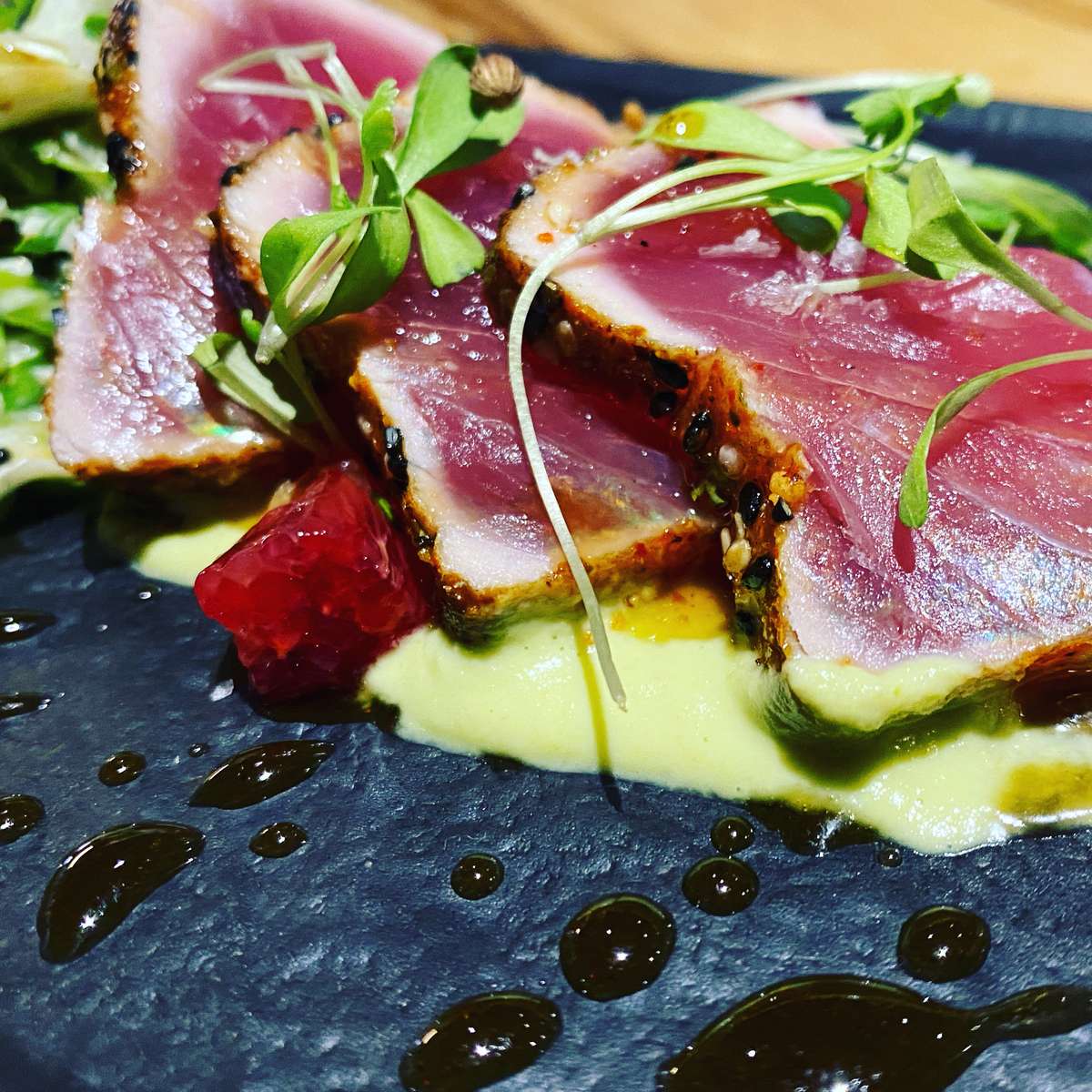 Sliced seared tuna with a pink centre, served over creamy green purée with fresh greens and a dark balsamic-style drizzle.
