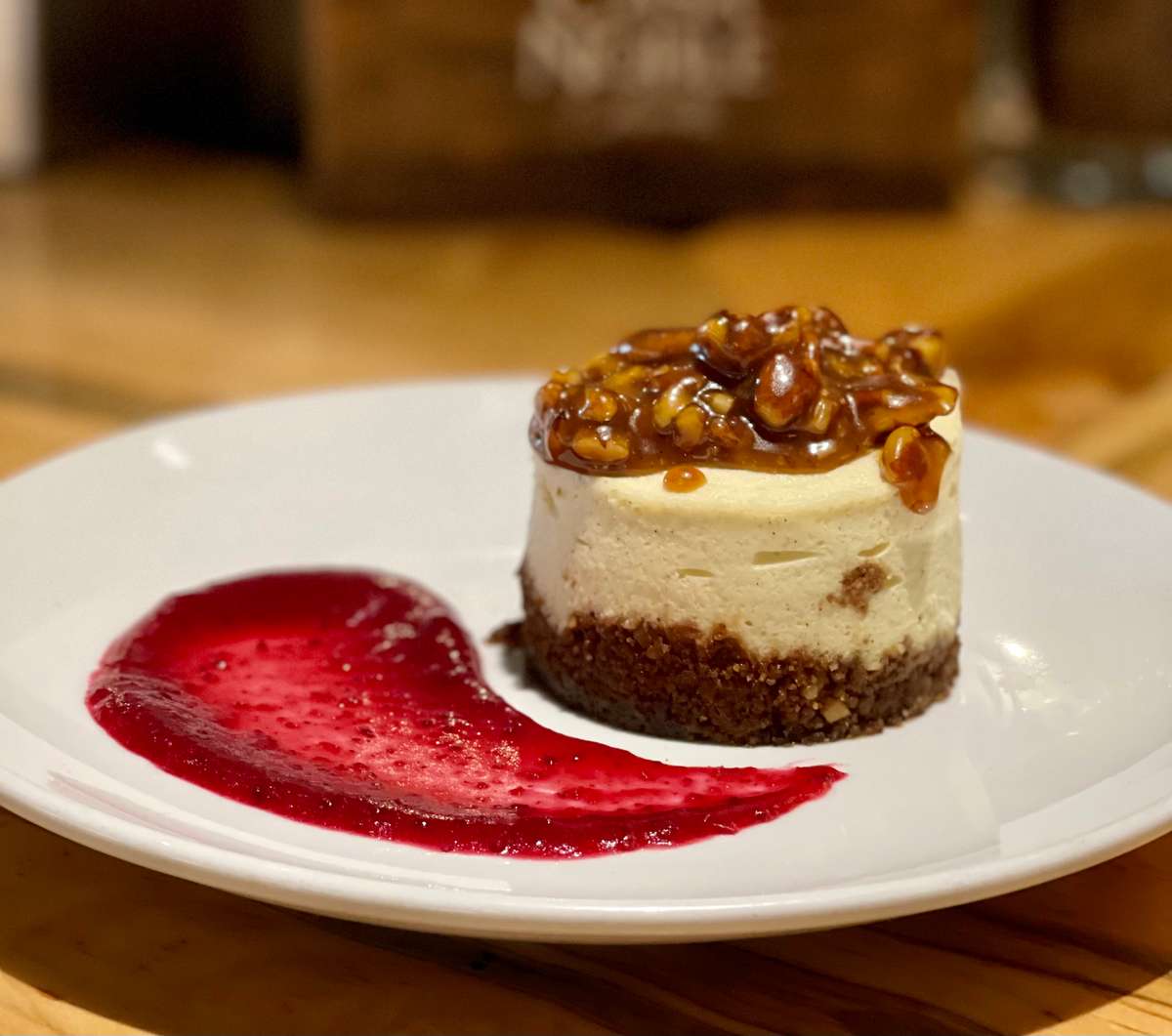 Individual cheesecake topped with caramelised nuts, served with a vibrant red berry sauce on a white plate.