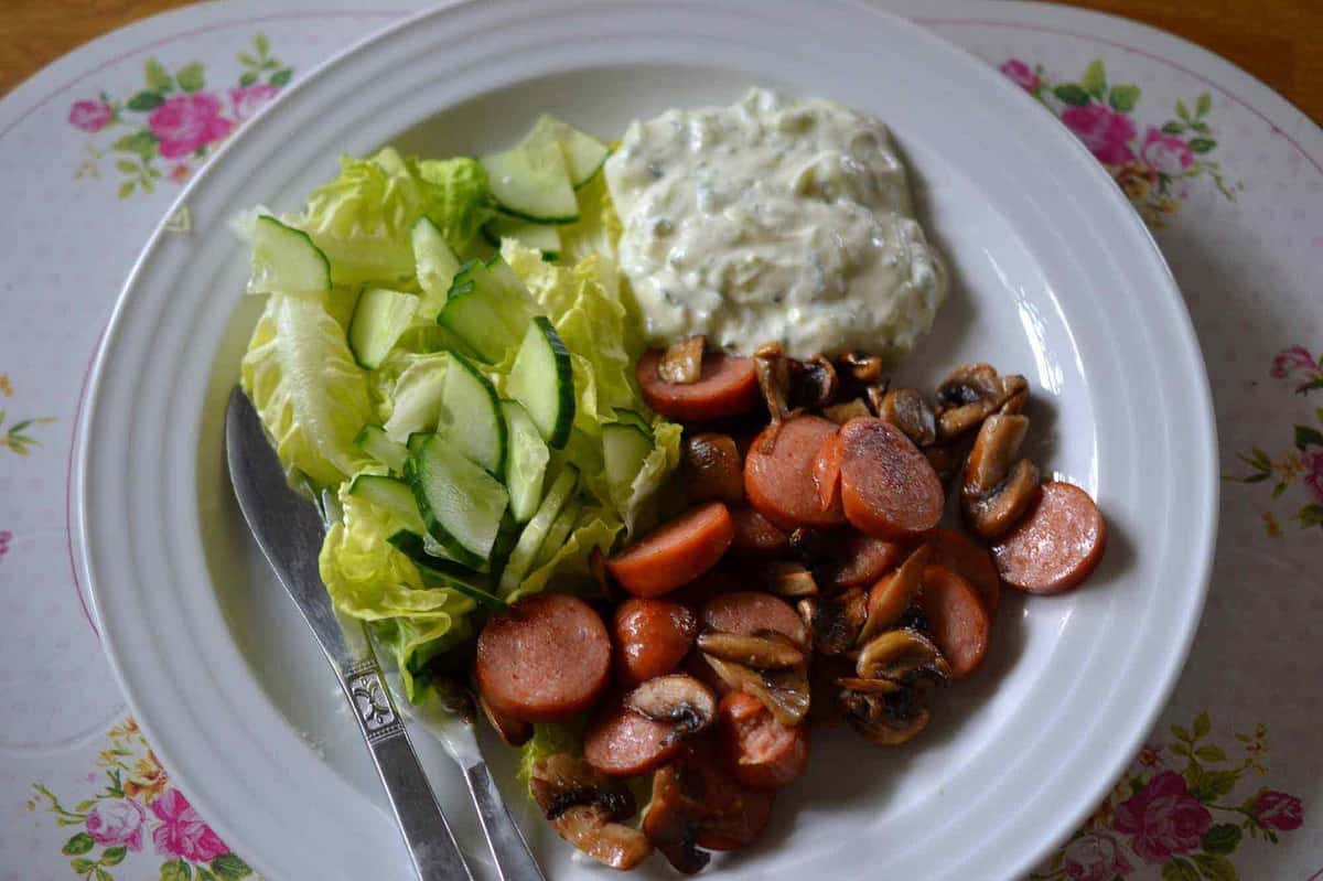 Grilled sausage slices with sautéed mushrooms, creamy potato salad, and a fresh cucumber-lettuce salad served on a floral plate—simple European-style home-cooked meal.