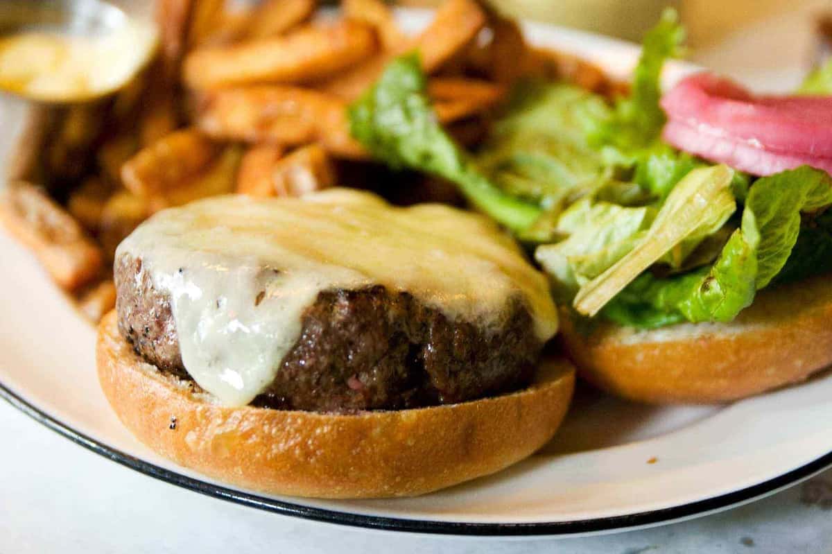 Juicy cheeseburger with melted white cheese on a toasted bun, served open-faced with fresh lettuce and pickled onions, alongside crispy seasoned fries and dipping sauce—classic American restaurant burger plate.