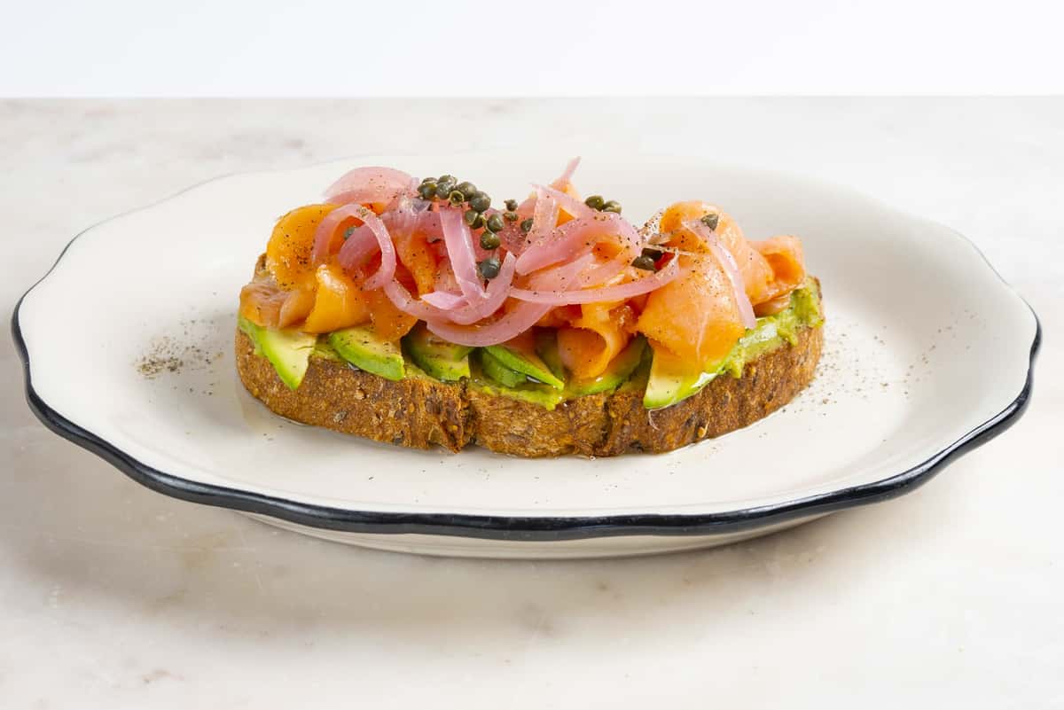Smoked salmon avocado toast topped with pickled red onions, fresh herbs, and creamy avocado on toasted artisan bread.