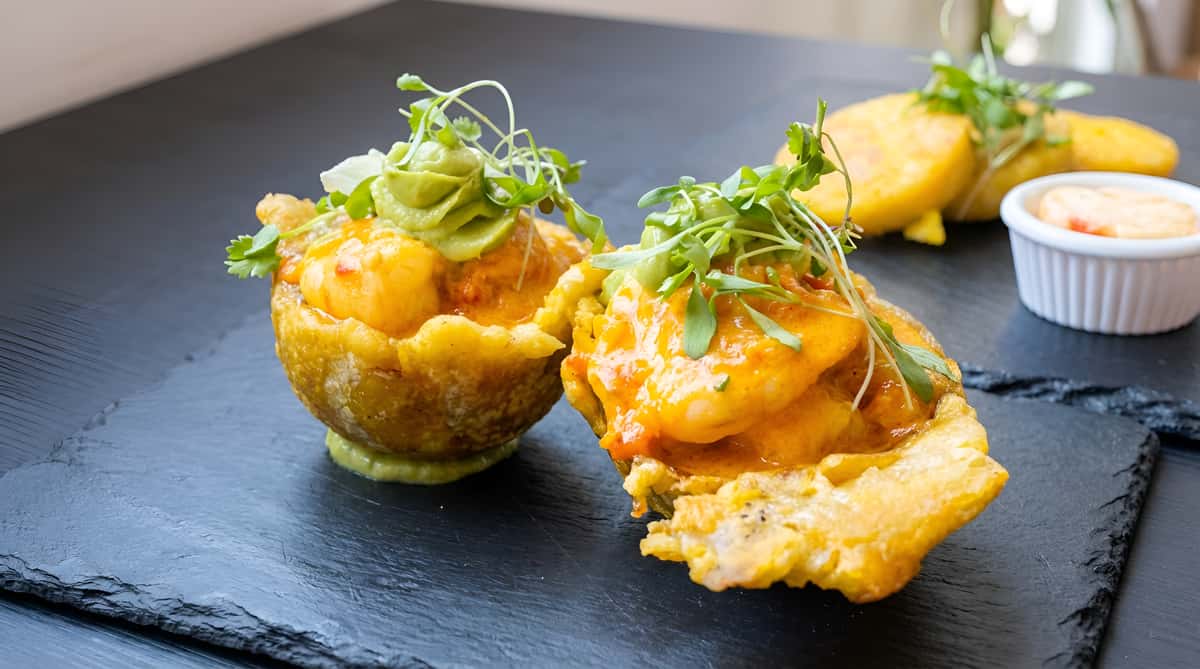 Two shrimp tostones topped with a creamy sauce and microgreens on a dark plate